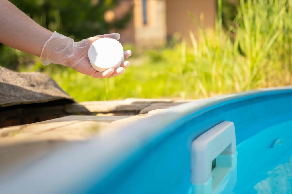 Female hand in rubber glove holds white chlorine pill over skimmer of swimming pool with blue water. Cleaning, disinfection of water in swimming pool.
